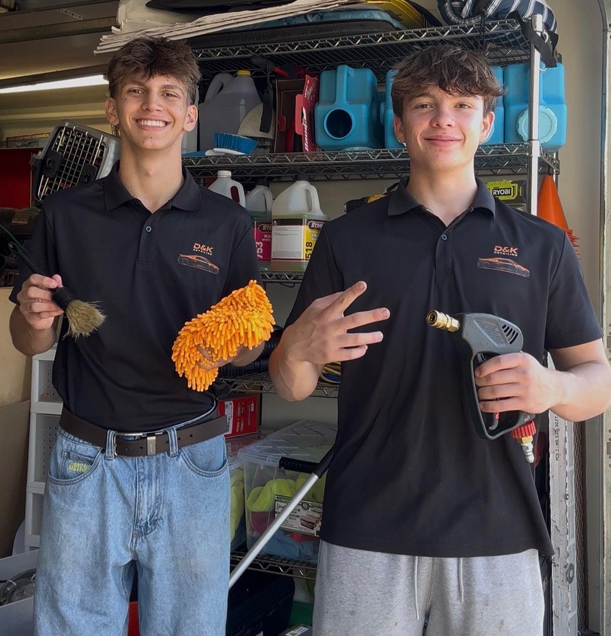 Two young workers in black polos holding cleaning supplies in front of a garage shelf filled with equipment