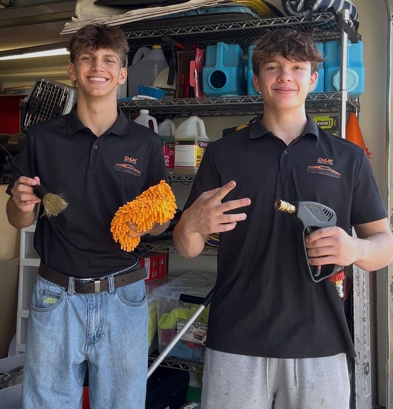 Two young workers in black polos holding cleaning supplies in front of a garage shelf filled with equipment