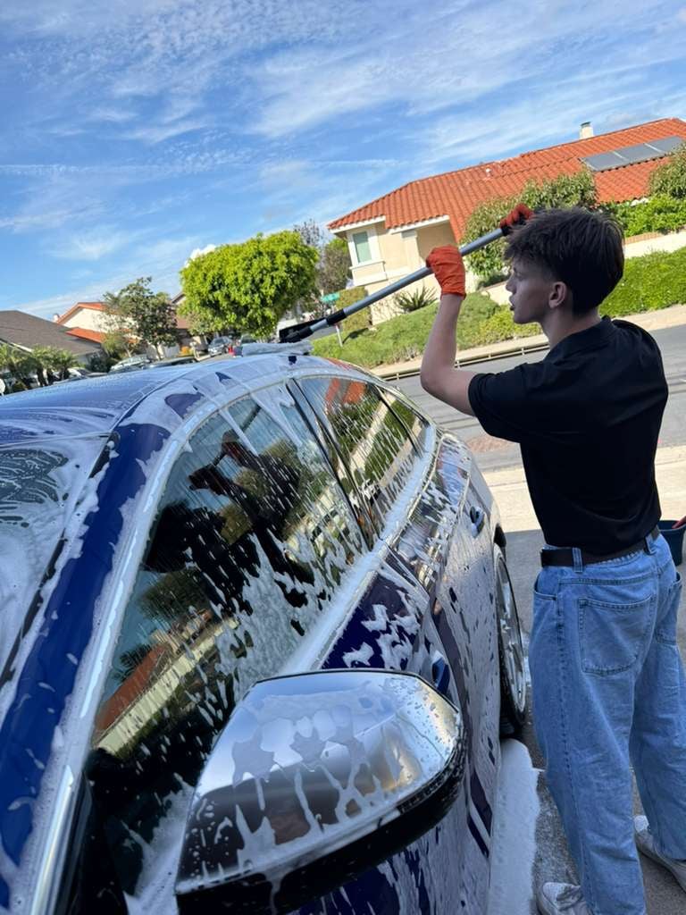 Man washing a blue car with a soapy sponge in a suburban driveway on a sunny day